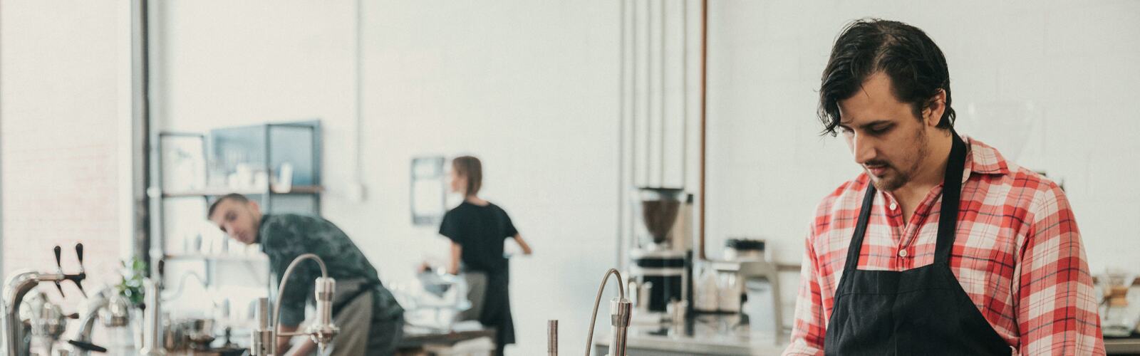 A café owner in an apron working at his counter while staff prepare drinks behind him