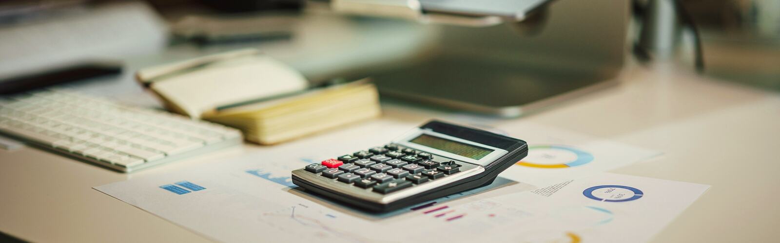 A calculator on printed budget charts beside a notebook and laptop on a desk