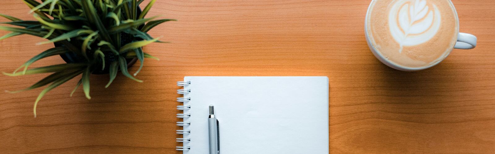 A blank notebook and pen beside a latte and a potted plant on a wooden table