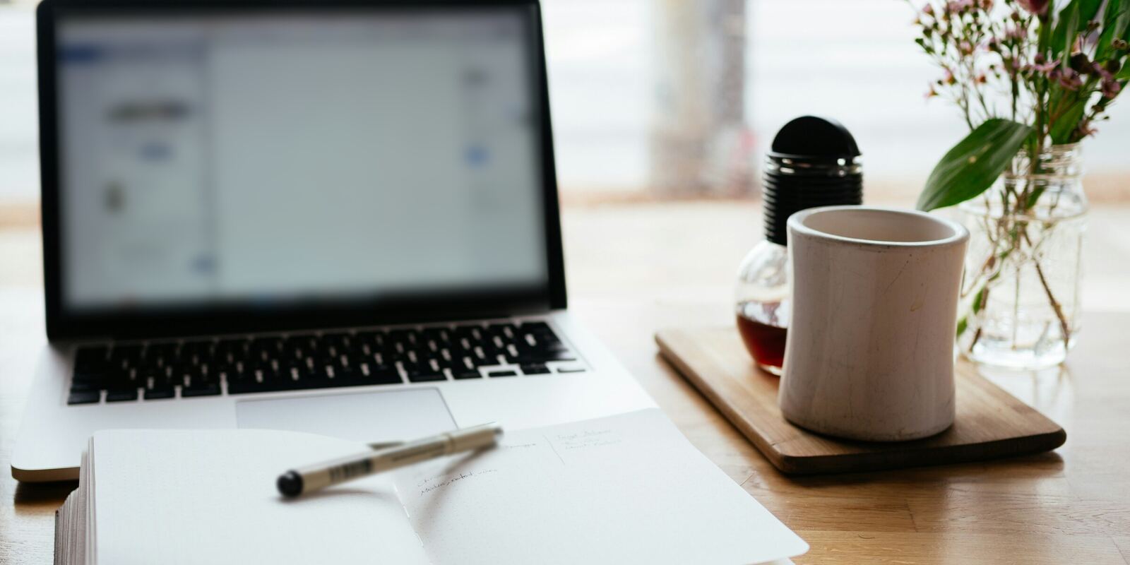 Un cuaderno abierto y una pluma junto a una laptop, una taza y un pequeño florero sobre una mesa de madera soleada
