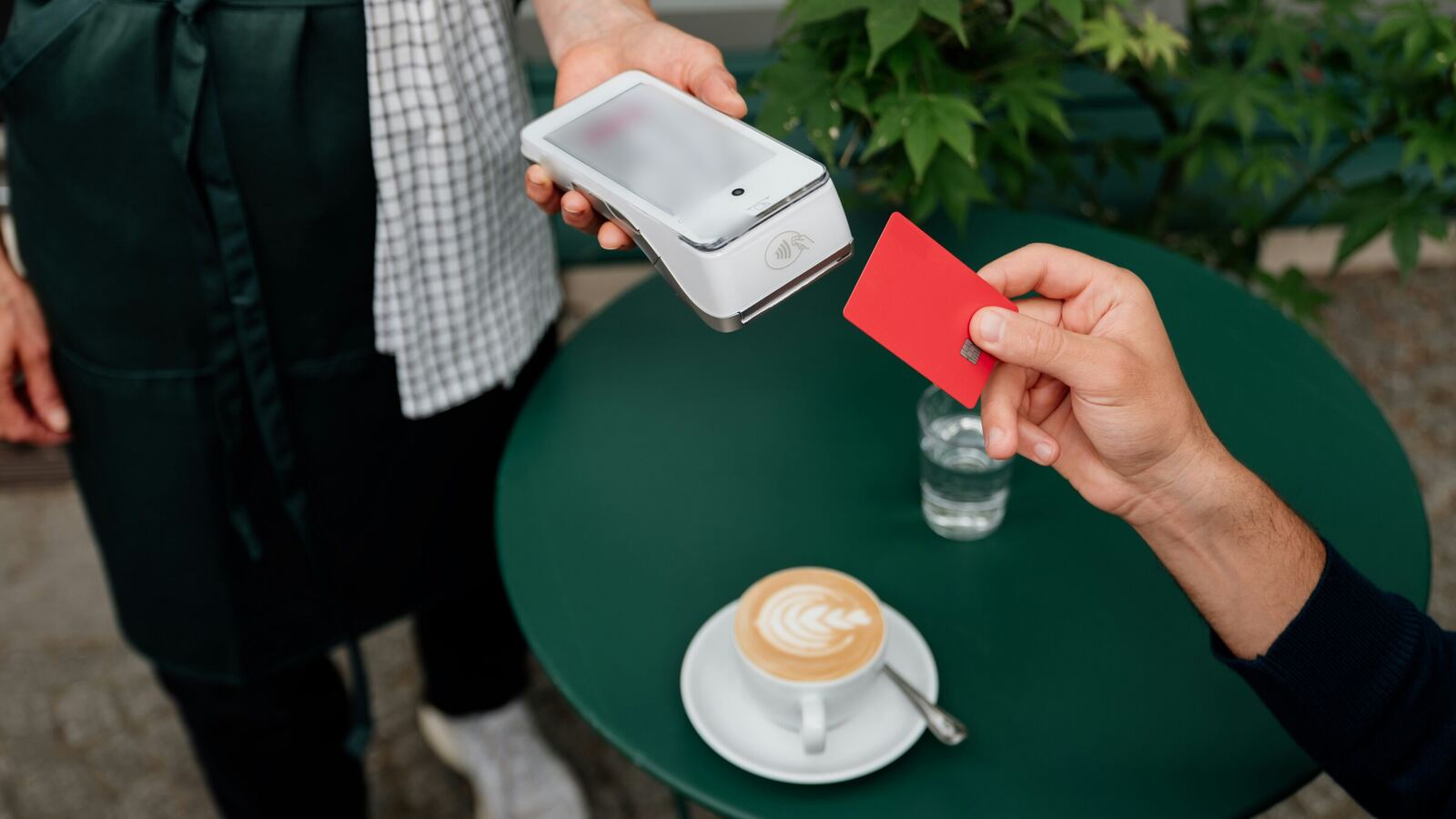 Un cliente acerca una tarjeta roja sin marcas a la terminal de pago que sostiene un mesero de café, junto a un capuchino sobre una mesa verde